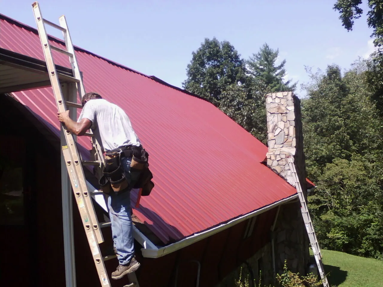 Roofer on a ladder beside a red metal roof and stone chimney — Soffit & Fascia Repair by Roofing Brothers in Letart